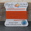 A spool of bright orange bead thread from Magnolia Bead Company, labeled Griffin 100% Natural Silk, set against a rustic wooden background.