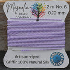 Spool of lavender thread with a label from Magnolia Bead Company on a wooden surface.