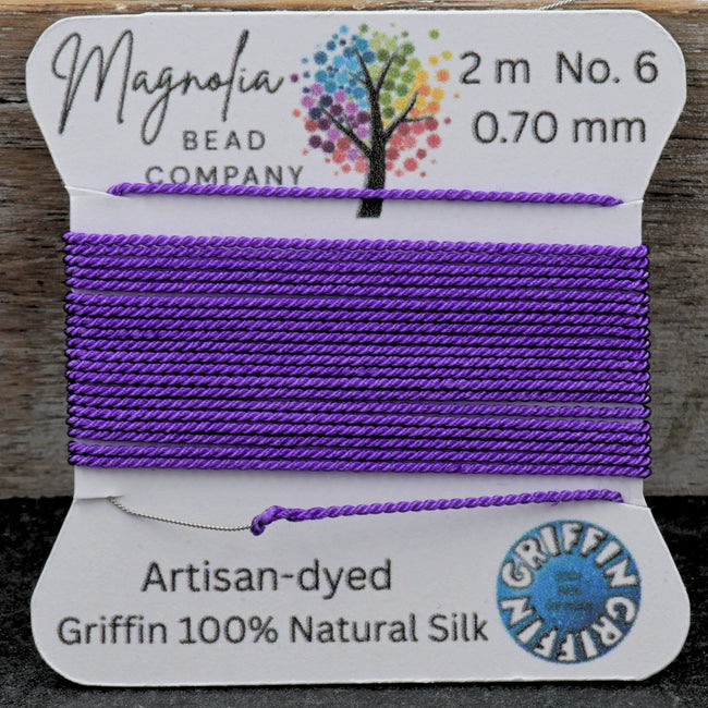 Spool of purple thread on a card with 'Magnolia Bead Company' branding, on a wooden surface. Hover Image