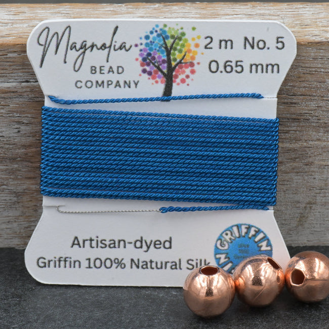 Blue thread on a card with 'Magnolia Bead Company' branding, next to three copper beads on a wooden surface. Hover Image