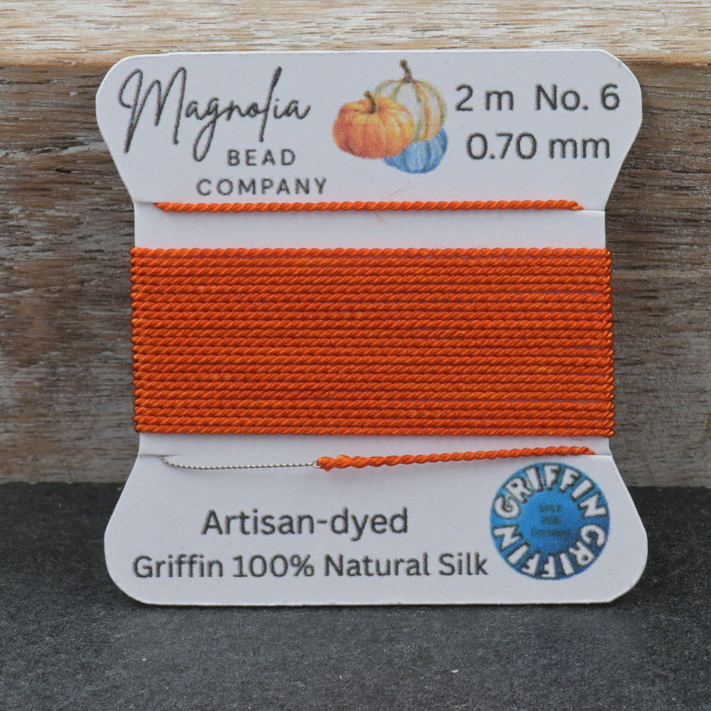 A spool of bright orange bead thread from Magnolia Bead Company, labeled Griffin 100% Natural Silk, set against a rustic wooden background.