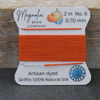 A spool of bright orange bead thread from Magnolia Bead Company, labeled Griffin 100% Natural Silk, set against a rustic wooden background.