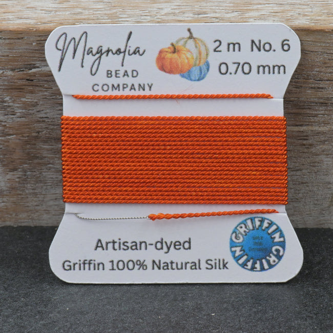 A spool of bright orange bead thread from Magnolia Bead Company, labeled Griffin 100% Natural Silk, set against a rustic wooden background. Hover Image