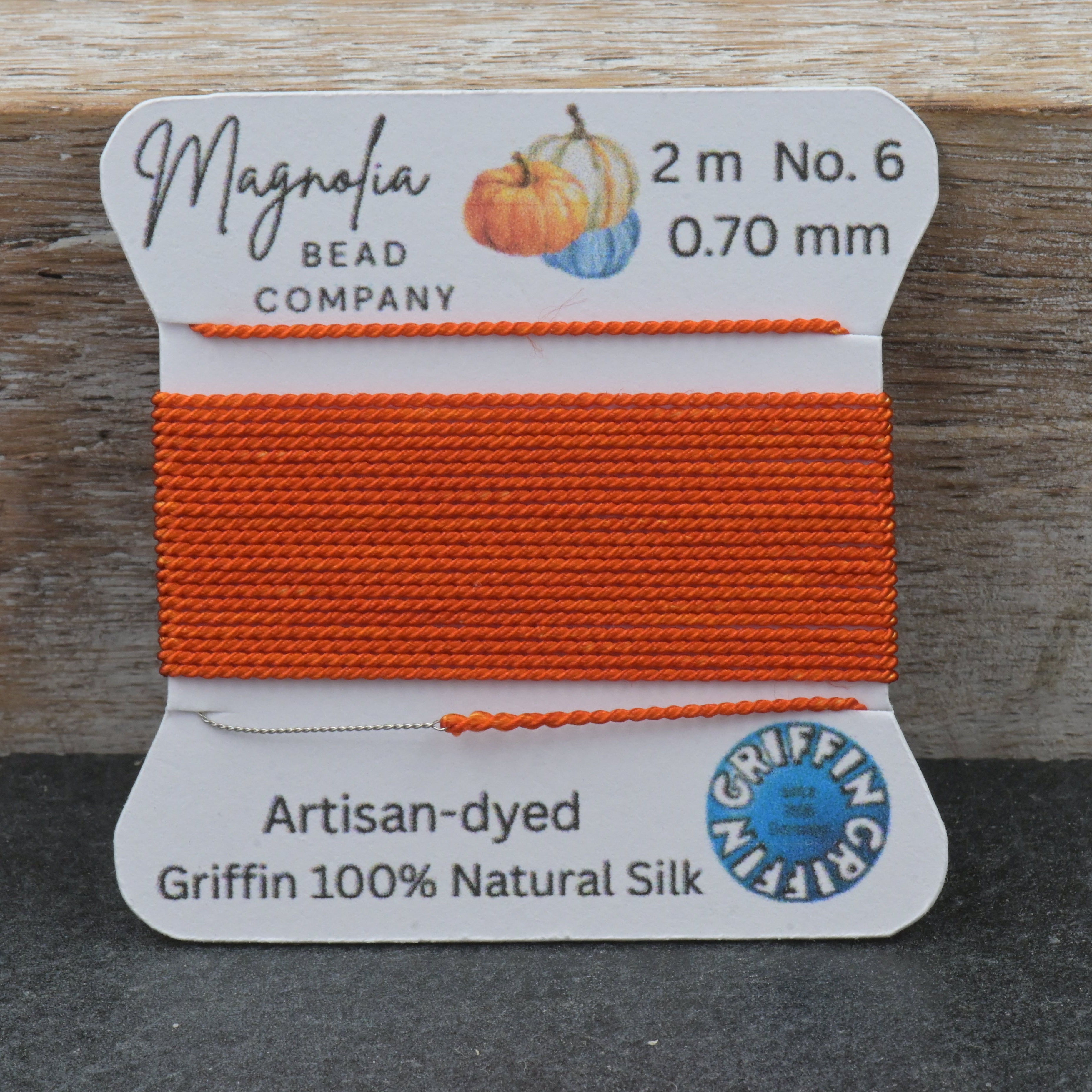 A spool of bright orange beading thread from the Magnolia Bead Company, Griffing Atlantis edition, displayed on a rustic wooden background.