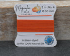 A spool of bright orange bead thread from the Magnolia Bead Company, labeled Artisan-dyed Griffin 100% Natural Silk, shown on a rustic wooden block against a dark gray surface.