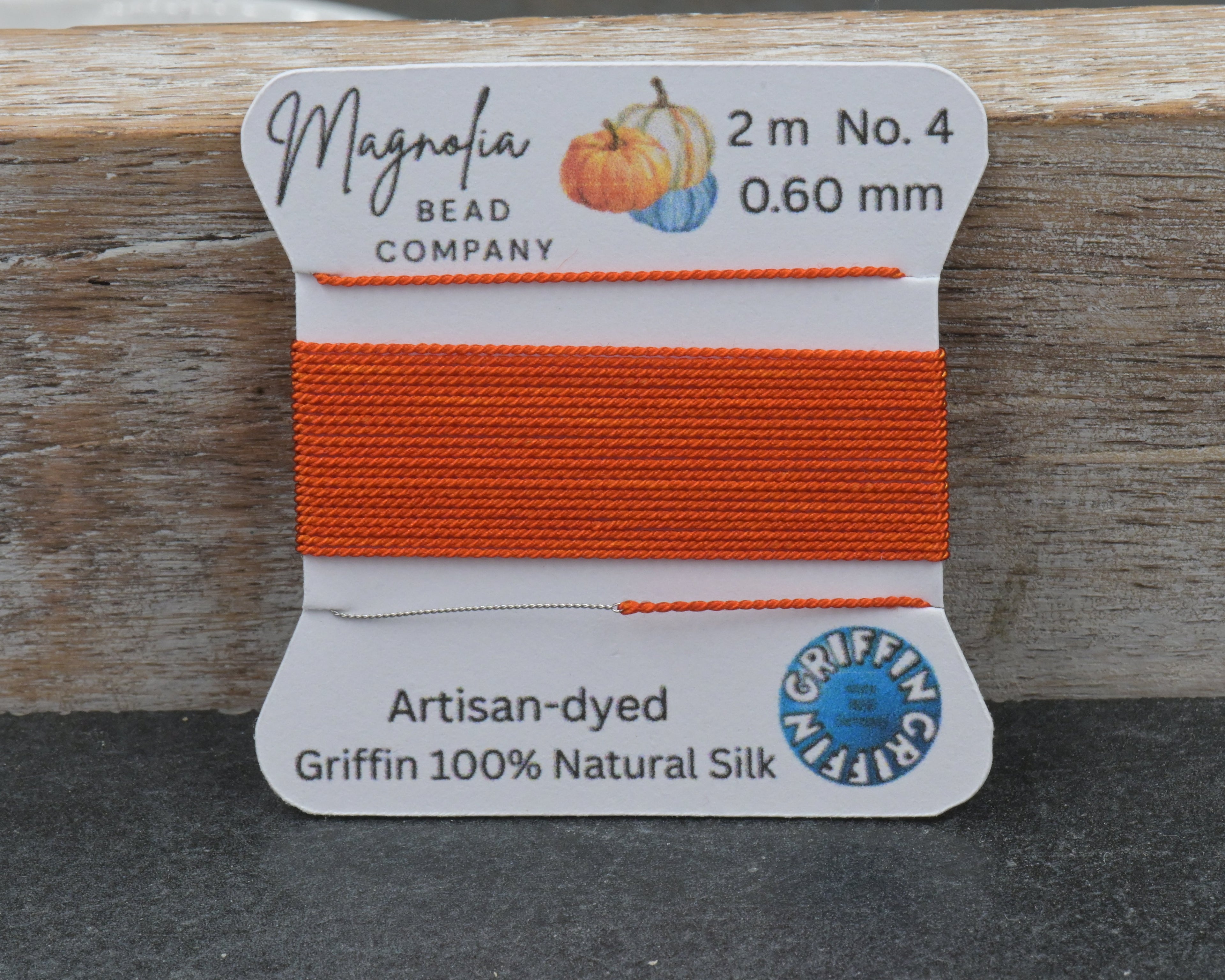 A spool of bright orange bead thread from the Magnolia Bead Company, labeled Artisan-dyed Griffin 100% Natural Silk, shown on a rustic wooden block against a dark gray surface.