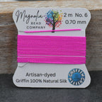 A spool of bright pink bead thread on a white Magnolia Bead Company card, labeled Artisan-dyed Griffin 100% Natural Silk, set against a rustic wooden background.