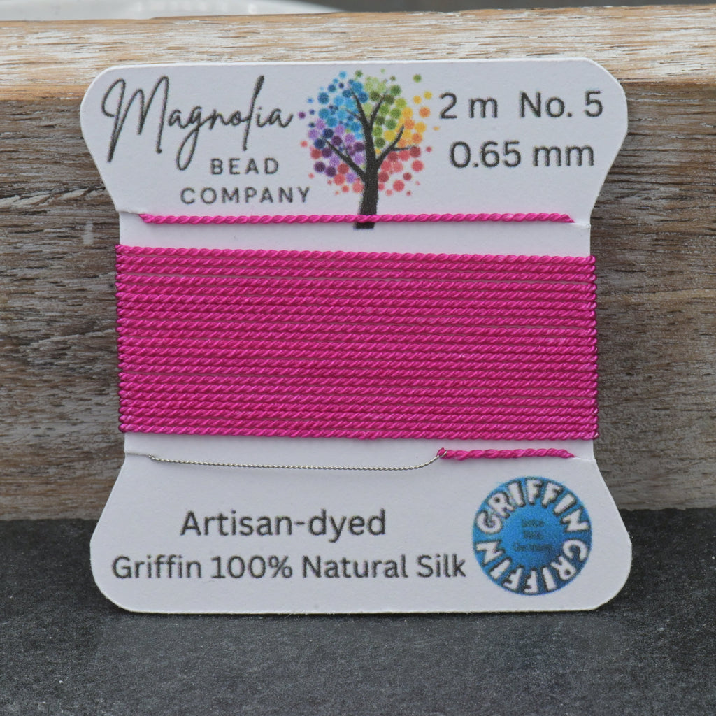 A spool of bright pink bead thread labeled 'Magnolia Bead Company' and 'Griffin 100% Natural Silk' resting on a dark gray surface with a rustic wooden background.