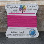 A spool of bright pink bead thread labeled 'Magnolia Bead Company' and 'Griffin 100% Natural Silk' resting on a dark gray surface with a rustic wooden background.