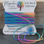 A spool of rainbow-colored artisan-tied silk cord beads from Magnolia Bead Company, with several loose ends draped in front, set against a rustic wooden background.