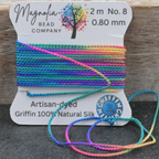 A spool of rainbow-colored artisan-tied silk cord beads from Magnolia Bead Company, with several loose ends draped in front, set against a rustic wooden background.