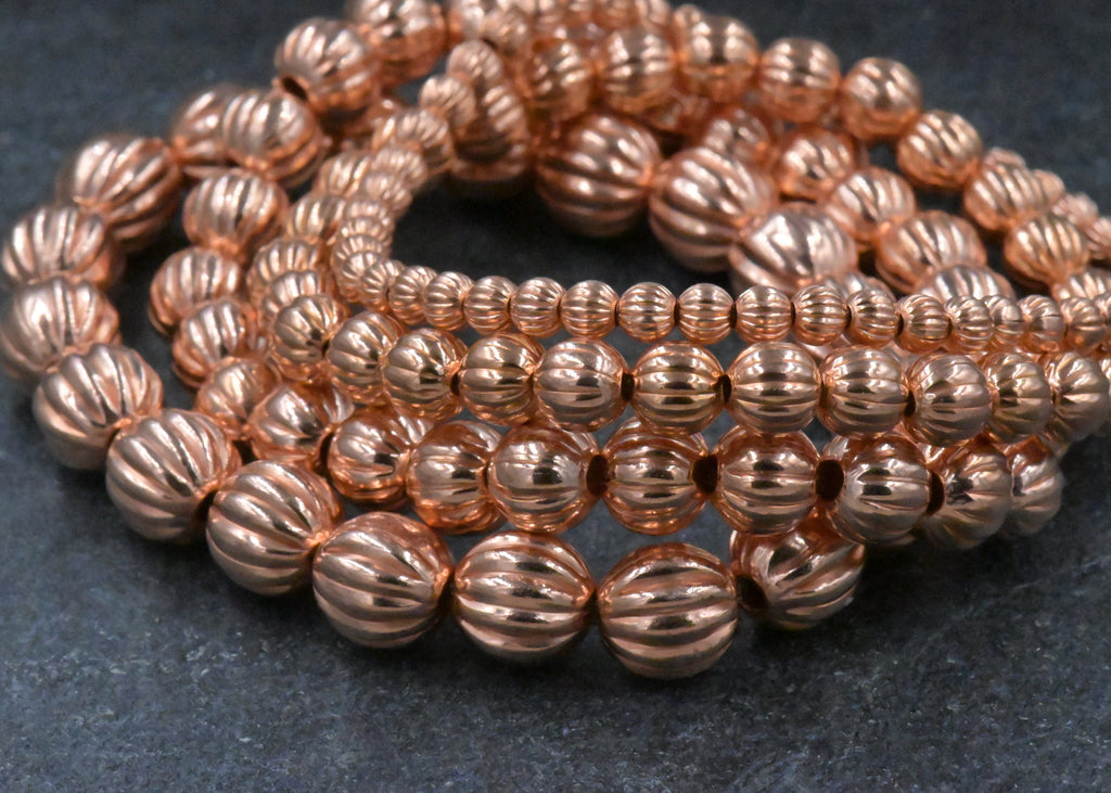 A close-up of multiple strands of shiny metallic rose-gold beads with a ribbed, textured surface, arranged on a dark gray textured background.